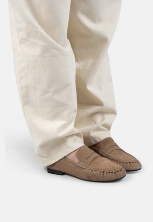 Persoon die beige suède loafers en loszittende gebroken witte broek draagt, staand op een witte achtergrond.