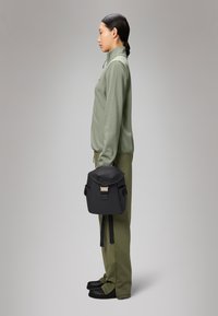 Mochila preta com fecho de rolo, fecho metálico e bolsos laterais, combinada com uma camiseta atlética verde clara e calças a condizer.