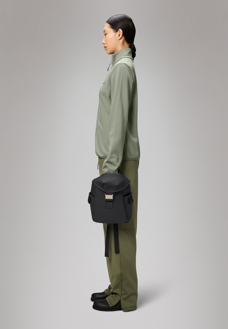 Mochila preta com fecho de rolo, fecho metálico e bolsos laterais, combinada com uma camiseta atlética verde clara e calças a condizer.