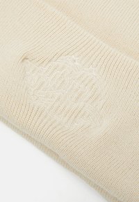 Krem beanie pletenice s teksturiranim uzorkom i utisnutim logotipom sa strane, izrađen od mekanog, laganog materijala.