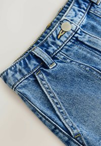 Jeans de mezclilla en azul claro con un acabado texturizado, que presentan un botón metálico, pasadores para cinturón y dos bolsillos delanteros. Las costuras son de un color en contraste.