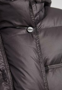 Primo piano di una giacca imbottita scura con cuciture visibili e un bottone a pressione nero tondo etichettato "JACKIT."