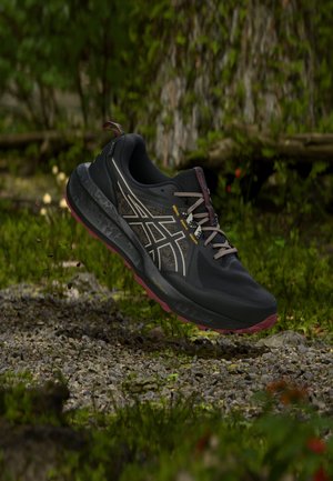 Chaussure de trail running noire et bordeaux avec des lacets gris suspendue au-dessus d'un sol forestier rocheux et des plantes vertes mousseuses.