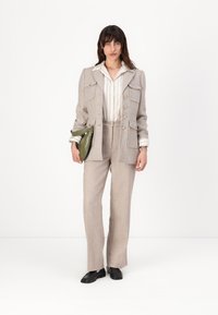 Veste de costume beige ajustée avec quatre poches, assortie à un pantalon. Chemise blanche à rayures en dessous. Chaussures noires et sac à main vert.