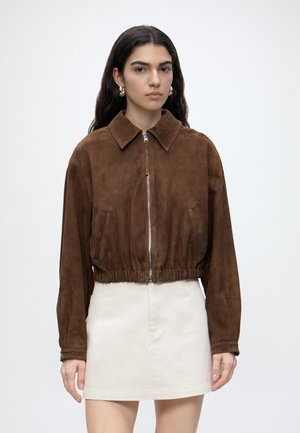 Woman with long dark hair wearing gold hoop earrings, a brown suede zip-up jacket, and a white mini skirt standing against a plain background.