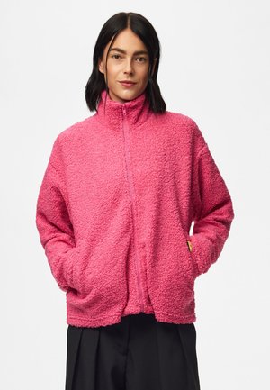 Femme aux cheveux foncés portant une veste zippée rose vif texturée, les mains dans les poches, sur un fond blanc uni.
