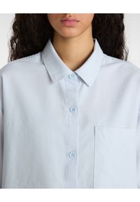 Camisa de botones azul claro hecha de tela suave. Cuenta con un cuello clásico, bolsillo frontal y cierre de botones. Diseño minimalista.