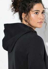 Felpa nera con cappuccio, caratterizzata da una consistenza morbida e un design semplice. Cuciture visibili e dettaglio con coulisse sul cappuccio.