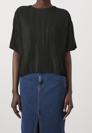 Personne portant un haut noir côtelé à manches courtes et une jupe en denim bleu avec une fente à l'avant, debout devant un fond uni.