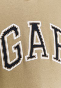 Gros plan sur les lettres "GAP" brodées en noir et blanc sur un tissu beige, montrant les détails de la broderie et la texture.