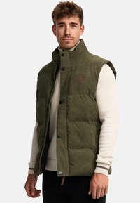 Vest matlasat de culoare verde măslin, cu guler înalt, având un patch cu logo, nasturi cu broșă și buzunare laterale. Purtați peste un pulover ușor.