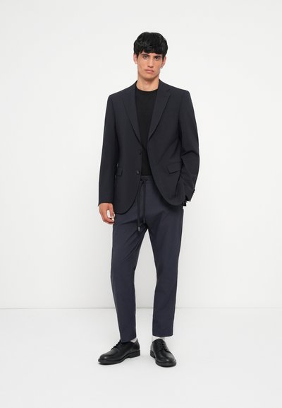 Blazer azul marino con dos botones, combinado con una camiseta negra y pantalones azul marino con cordón. Zapatos negros completan el atuendo.