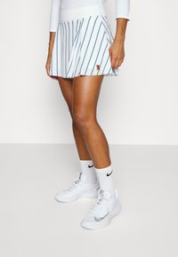 Falda de tenis blanca con pliegues y rayas verticales en tono verde azulado, con detalle de logo bordado, combinada con zapatillas deportivas blancas y calcetines hasta la mitad de la pantorrilla.