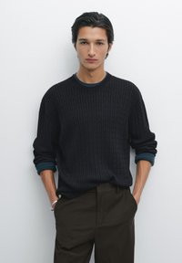Schwarzer Strickpullover mit geripptem Muster und runder Halslinie, kombiniert mit dunklen Olivenhosen. Blauer Akzent an den Ärmelbündchen.