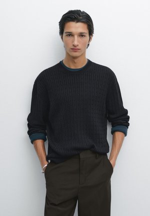 Schwarzer Strickpullover mit geripptem Muster und runder Halslinie, kombiniert mit dunklen Olivenhosen. Blauer Akzent an den Ärmelbündchen.