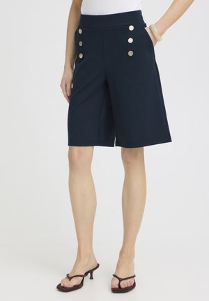 Nő tengerészkék térdig érő rövidnadrágot visel hat arany gombbal, fehér felsőt, bézs karperecet és sötét magas sarkú szandált, egyik kezét a zsebében tartva áll.