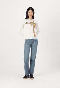 Witte sweater met gouden pailletten grafisch ontwerp van een strik en boog. Gepaard met lichtblauwe jeans en zwarte sneakers met een beige zool.