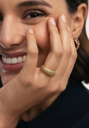 Anillo de oro adornado con piedras multicolores, que muestra un diseño liso y curvado. La mano del modelo toca suavemente su rostro, destacando el anillo.
