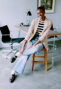 Blazer beige sobre una camiseta de rayas en blanco y negro, combinado con jeans acampanados de un azul claro y mocasines oscuros con un acento dorado. Suelo y silla lisos.