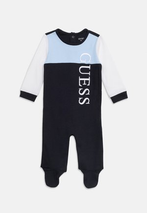 Barboteuse bébé manches longues avec pieds, panneau bleu sur la poitrine, manches blanches, poignets noirs et logo vertical "GUESS" sur le corps bleu marine.
