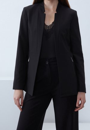 Femme portant un blazer noir par-dessus un haut noir bordé de dentelle et un pantalon noir assorti, debout les mains détendues le long du corps.