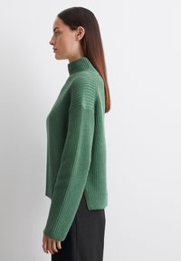 Grüner Strickpullover mit hohem Kragen, gerippter Textur, seitlichen Schlitzen und überschnittenen Schultern, kombiniert mit einem schwarzen Rock.