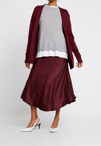 Burgunderfarbener Cardigan über einem grauen Pullover, einer weißen Bluse und einem glänzenden burgunderfarbenen Rock. Weiße High-Heels. Glatte Texturen, gestaffelter Look.
