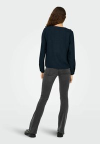 Navy textuur langslavige top met een vierkante halslijn, gedragen met grijze flared jeans en zwarte schoenen. Achteraanzicht benadrukt de stofdetails van het shirt.