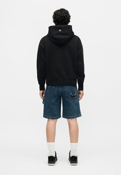 Άνδρας με μαύρη φούτερ με κουκούλα και μπλε τζιν σορτς στέκεται γυρισμένος με την πλάτη σε απλό φόντο.