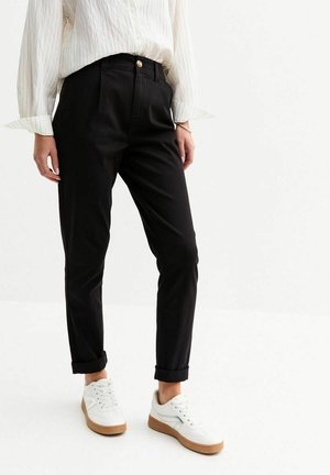 Personne portant un pantalon noir taille haute fuselé avec revers roulés, un chemisier blanc à fines rayures et des baskets blanches avec des semelles en caoutchouc, debout sur un sol blanc.