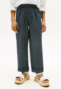 Pantaloni a gamba larga a righe blu e bianche con pieghe frontali, vita elastica e un tessuto strutturato, abbinati a sandali con plateau bianchi.