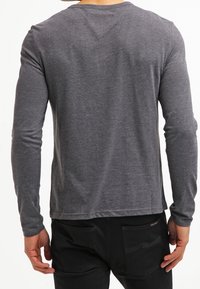 Mężczyzna w dopasowanej, szarej koszulce z długim rękawem i czarnych spodniach, odwrócony tyłem z rękami luźno opuszczonymi wzdłuż ciała.