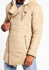 Persona che indossa una giacca trapuntata beige con cappuccio, tasche con zip e un maglione sotto, in posa con una mano vicino alla zip.
