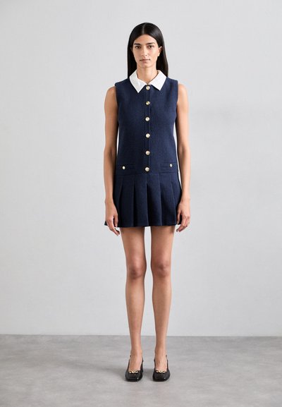 Robe sans manches marine avec un col blanc, des boutons dorés et une jupe plissée. Dispose de deux poches avant et d'un tissu texturé.