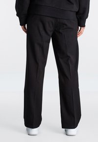 Schwarze weit geschnittene Hosen aus glattem Stoff, mit einem schlichten Design und subtilen Falten, wobei unten weiße Turnschuhe sichtbar sind.