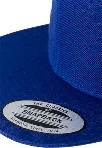 Blå snapback-kaps med et struktureret stof, flad skygge og et rundt logo-patch med teksten "SNAPBACK" samt mærkedetaljer.