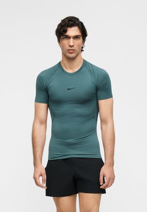 Mężczyzna ubrany w dopasowaną, krótką koszulkę Nike w kolorze morskim i czarne krótkie spodenki, stojący na jednolitym jasnym tle.