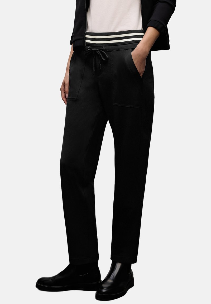 Schwarze Hose mit Kordelzugbund und gestreiftem elastischem Band. Glatter Stoff mit Seitentaschen und geraden Beinen, getragen mit schwarzen Schuhen.