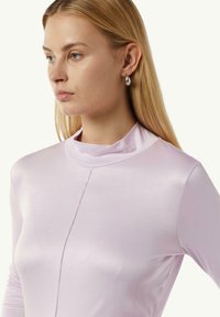 Helles rosa Langarmoberteil mit hohem Kragen, glatter Satin-Textur und Vordernahtdetails, kombiniert mit einem schlichten silbernen Creolenohrring.