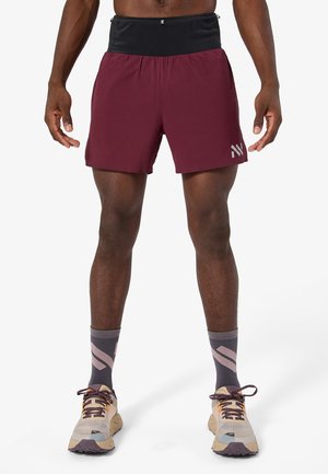 Hombre vistiendo pantalones deportivos color granate con cintura negra, calcetines grises y rosas, y zapatos para correr beige, de pie contra un fondo blanco.