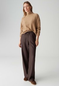 Pull en maille beige à col montant, associé à un pantalon ample marron foncé avec une taille élastique et un cordon de serrage. Des chaussures marron complètent la tenue.