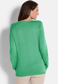 Frau mit langen braunen Haaren, die einen leuchtend grünen Langarmpullover und beige Hosen trägt, von hinten schaut und lächelnd zur Seite blickt.