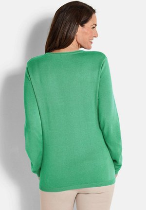 Frau mit langen braunen Haaren, die einen leuchtend grünen Langarmpullover und beige Hosen trägt, von hinten schaut und lächelnd zur Seite blickt.