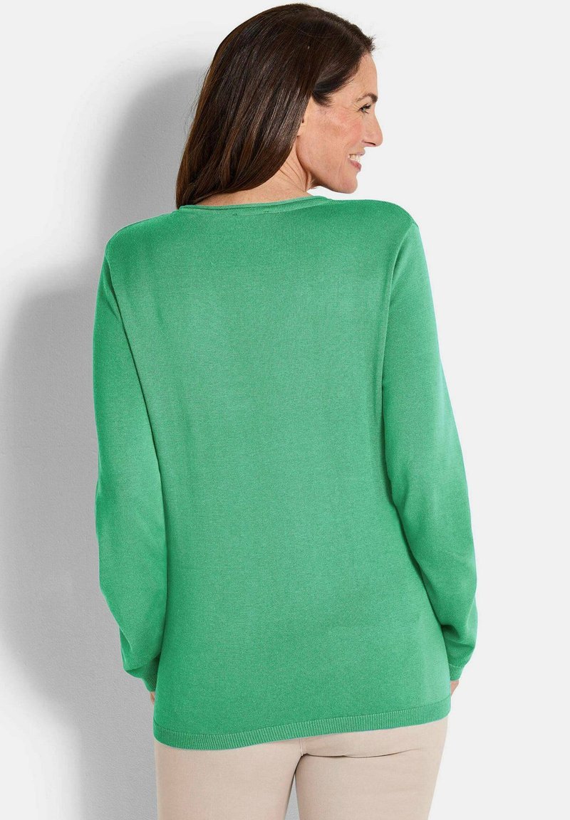 Frau mit langen braunen Haaren, die einen leuchtend grünen Langarmpullover und beige Hosen trägt, von hinten schaut und lächelnd zur Seite blickt.
