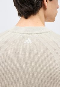 Άτομο που φαίνεται από πίσω να φορεί ένα μπεζ, υφασμάτινο πλεκτό πουλόβερ με μικρό λευκό λογότυπο Adidas στο κέντρο κάτω από το λαιμό.