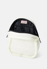Mochila blanca acolchada con una apertura con cremallera, que cuenta con un interior negro con bolsillos de organización. La etiqueta de la marca es visible en el interior.