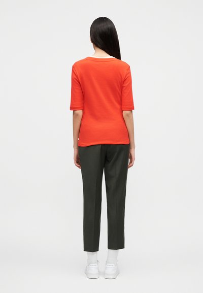 Γυναίκα που στέκεται με την πλάτη προς τα εμπρός φοράει πορτοκαλί κοντομάνικη μπλούζα, σκούρο γκρι παντελόνια, λευκές κάλτσες και λευκά αθλητικά παπούτσια σε απλό φόντο.