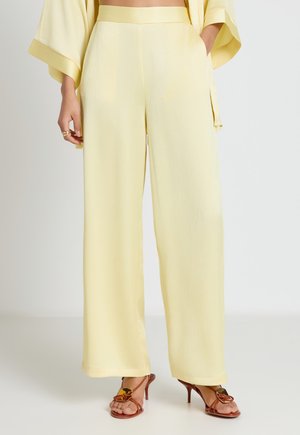 Woman wearing high-waist, wide-leg pale yellow pants with side pockets and brown open-toe heeled sandals.