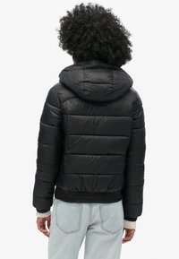 Sort dunjakke med hætte, med quiltede sektioner, elastiske manchetter og en tætsiddende talje. Båret over lyseblå jeans.