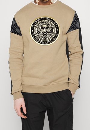 Beżowa bluza z czarnym wzorzystym materiałem na rękawach oraz okrągłą emblematem z pszczołą i napisem "Glorious Gangsta" na piersi.